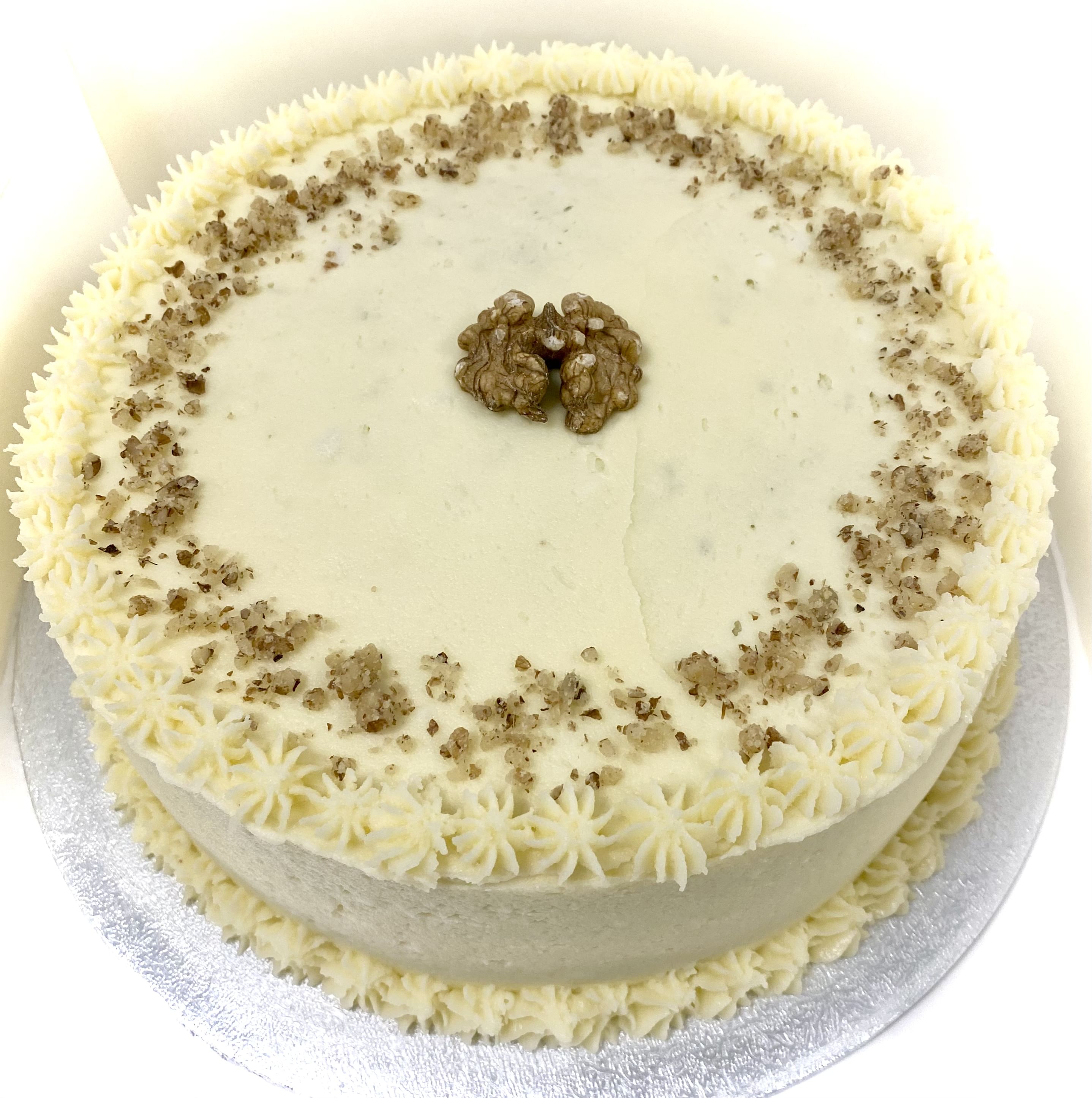 A round cake with creamy white frosting, decorated with a ring of finely chopped nuts on top. A single walnut sits in the center. The cake is placed on a silver board. The frosting is piped in small rosettes around the top edge.
